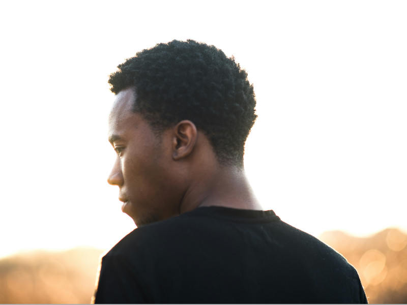 A Black man stands, back to the camera, his head is turned to the side, features partially obscured by light in the background